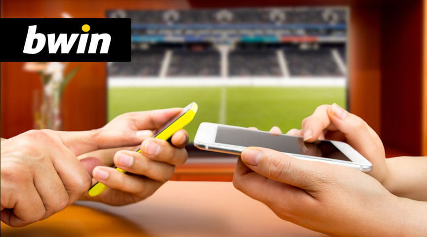 Le mani di due scommettitori che piazzano una giocata sui loro smartphone. Sullo sfondo il logo di bwin e lo schermo di una tv che trasmette una partita di calcio